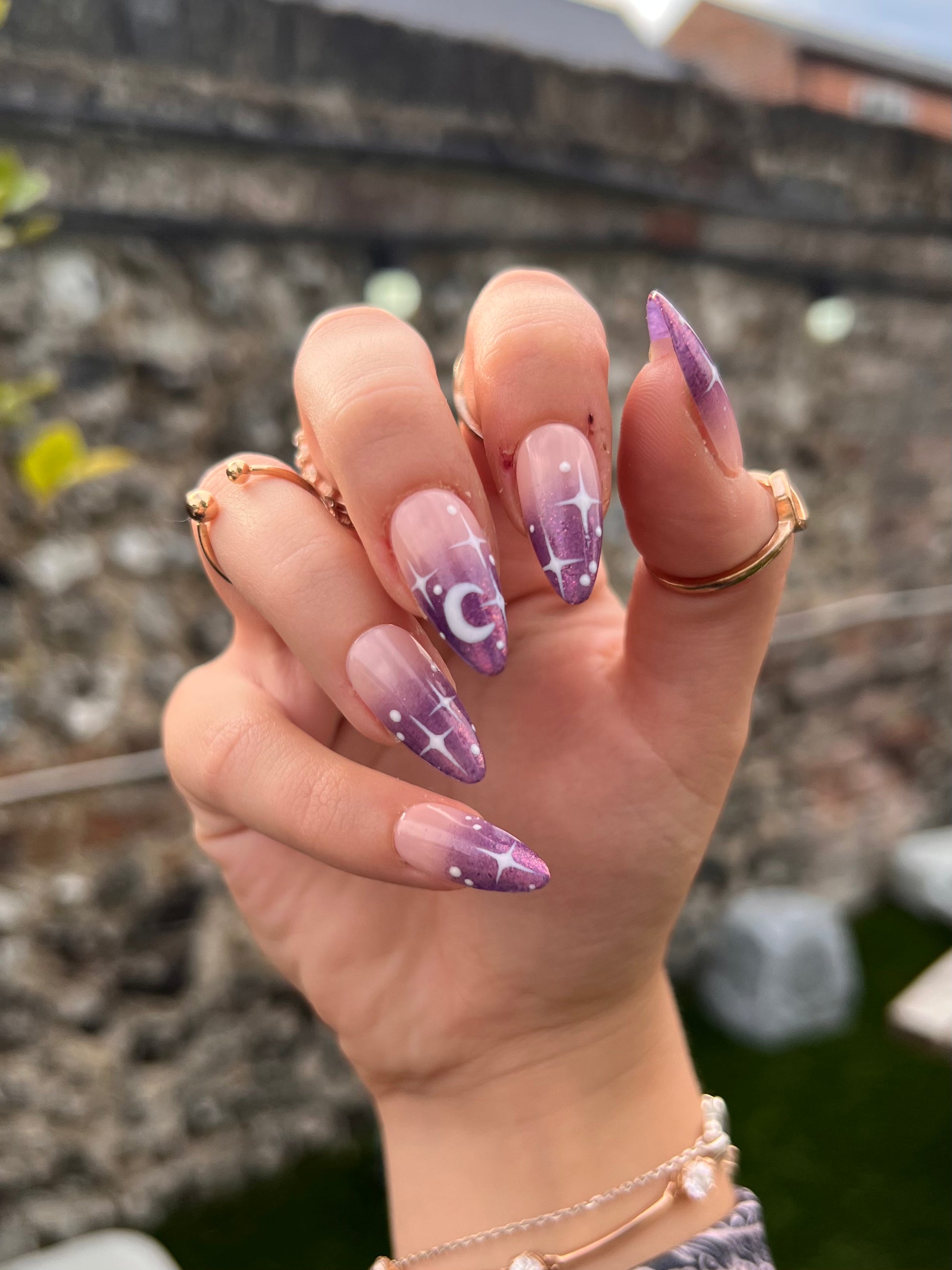 Mid-length natural almond fake nails with celestial stars and moon design. Glitter purple ombre over a nude base.